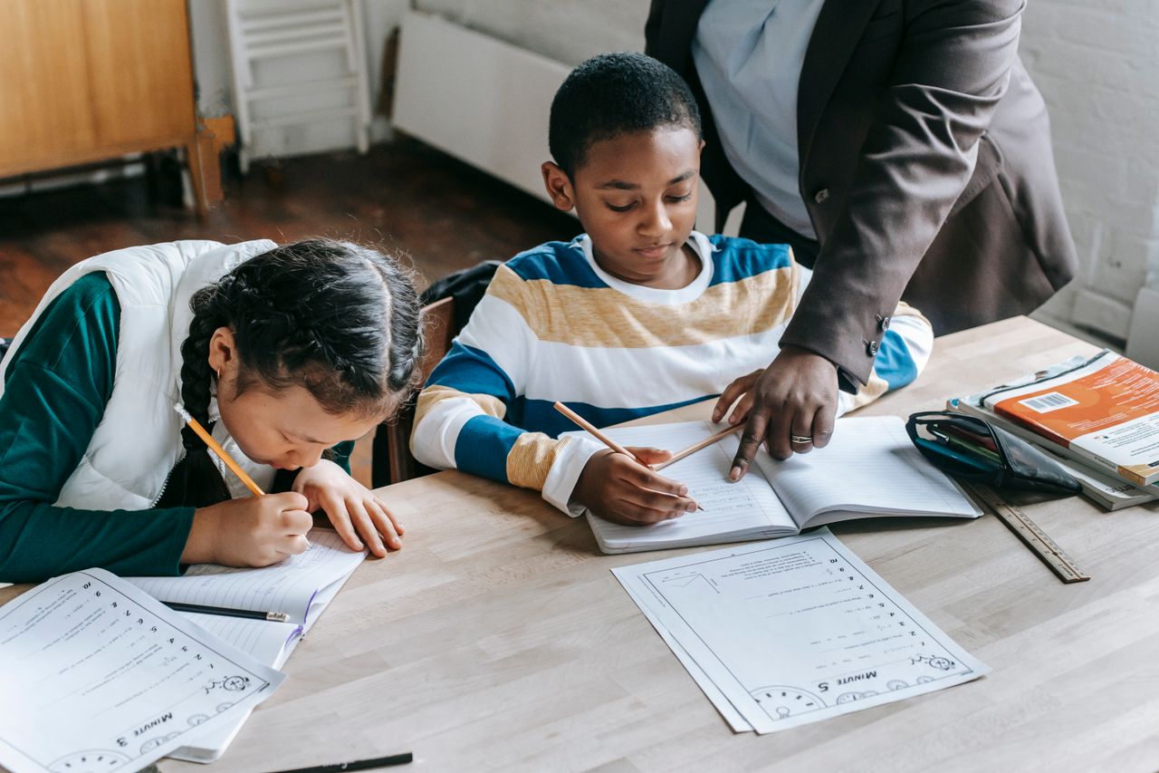 hero-gallery-02 High angle of crop unrecognizable black female teacher explaining task to focused little schoolboy sitting at desk near attentive Asian classmate writing in notebook